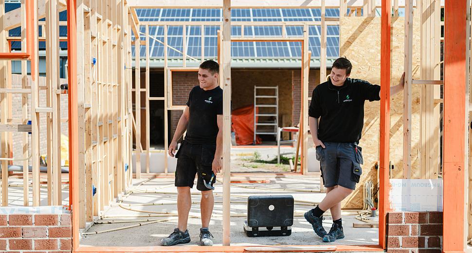 Twee monteurs in werkkleding leunen ontspannen tegen een houten frame van een nieuwbouwproject, met bakstenen onderbouw en zicht op zonnepanelen in de achtergrond.