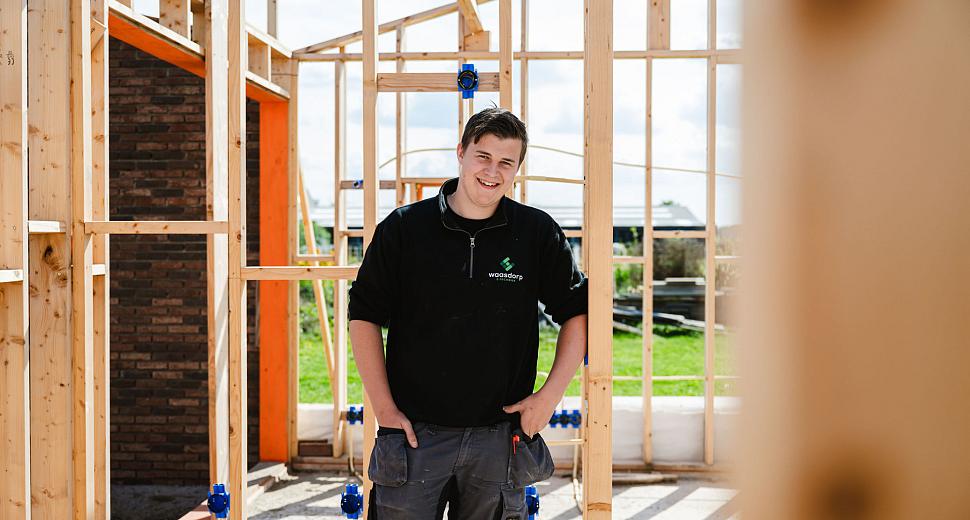 Een installateur in werkkleding poseert glimlachend binnen een houten frame van een nieuwbouwproject, met blauwe inbouwdozen en een zonnige tuin op de achtergrond.