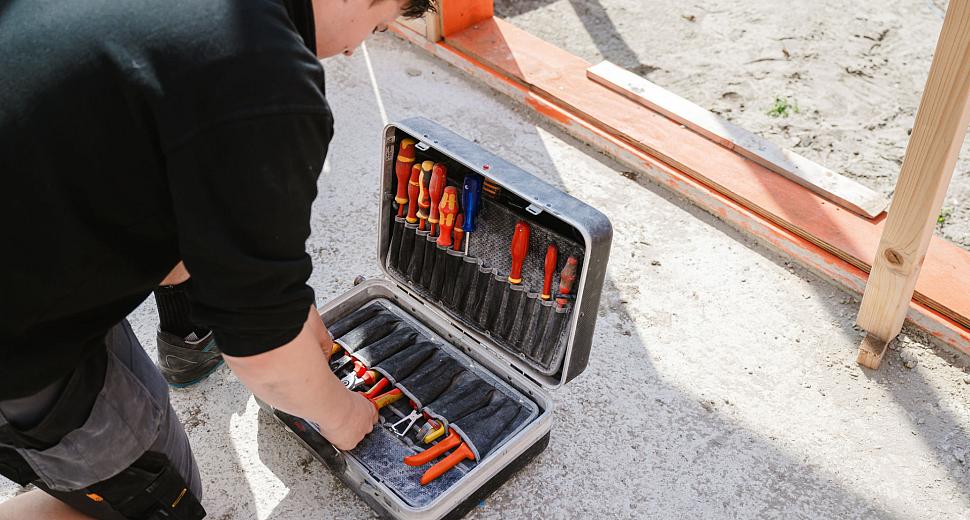 Monteur knielt naast een geopende gereedschapskoffer gevuld met handgereedschap zoals schroevendraaiers en tangen, op een bouwplaats met houten frameconstructie op de achtergrond.