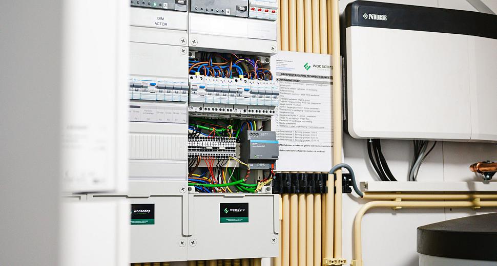 Een technische ruimte met een geavanceerde schakelkast gevuld met bedrading, automaten en modules, omringd door verwarmingsbuizen en een ventilatiesysteem.