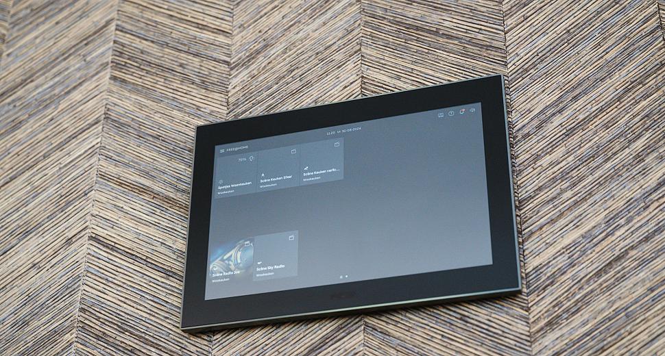 Wandgemonteerd touchscreen voor domotica-bediening met een strak ontwerp, geïnstalleerd tegen een decoratieve wand met een visgraatstructuur.
