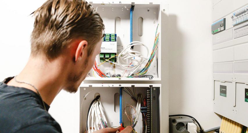 Technicus werkt aan een open schakelkast met kleurrijke bedrading en geavanceerde apparatuur, naast een paneel met moderne elektrische systemen.