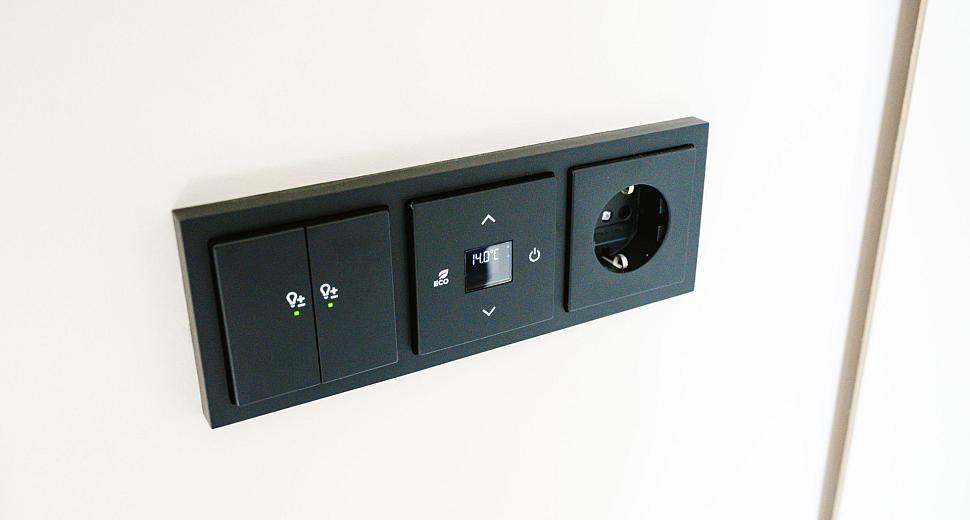 Zwart wandpaneel met geïntegreerde lichtschakelaars, een digitale thermostaatweergave en een stopcontact, gemonteerd op een witte muur.