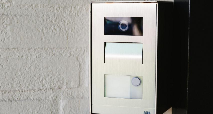  RVS deurbelpaneel met geïntegreerde camera, microfoon en belknop, gemonteerd op een witte bakstenen muur naast een zwarte deurstijl.