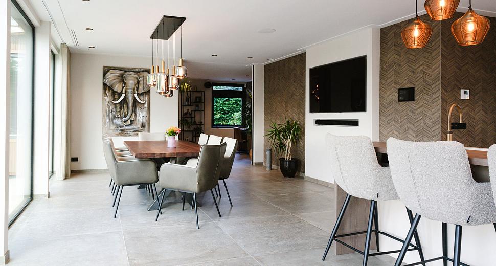 Een moderne eetkamer met een grote houten eettafel, omringd door gestoffeerde stoelen, en hanglampen boven de tafel. Links glazen schuifdeuren met uitzicht op buiten.