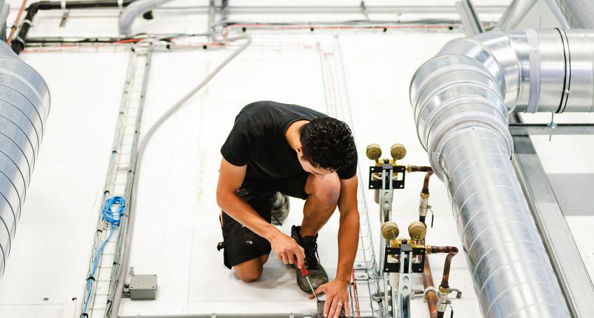  Monteur aan het werk op een technische installatie, bezig met het verbinden van leidingen en kabels tussen ventilatiesystemen en elektriciteitskabels.