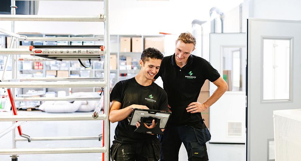 Twee technici in Waasdorp-kleding bekijken samen een tablet in een werkplaats met ladders, gereedschap en technische apparatuur op de achtergrond.