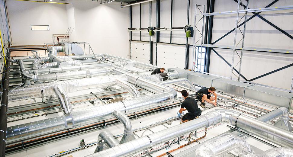 Technici werken aan de installatie van een geavanceerd ventilatiesysteem met metalen luchtkanalen in een moderne industriële ruimte met hoge plafonds.