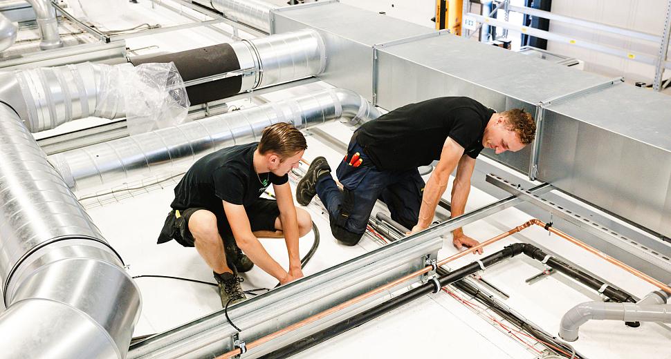 Twee technici werken aan leidingen en kabels op een technische werkplek met ventilatiekanalen en kabelgoten in een industriële omgeving.