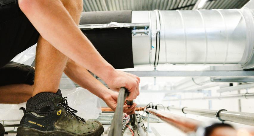 Technicus knielt op een verhoging in een industriële ruimte en werkt aan kabels, omringd door ventilatiekanalen en een metalen plafondconstructie.