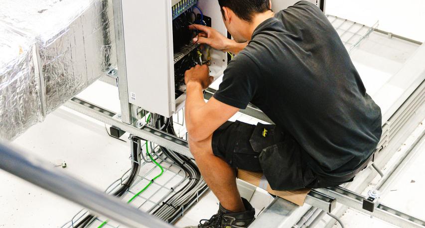 Technicus zit op een verhoging en werkt aan een open schakelkast in een technische ruimte met zichtbare ventilatiekanalen en kabelgoten.