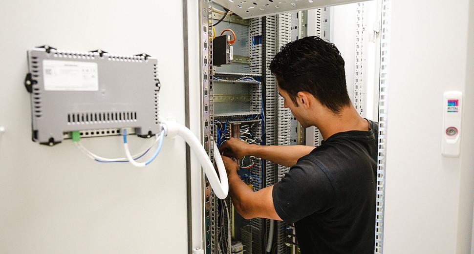 Technicus werkt aan de bedrading in een open schakelkast met complexe elektronische componenten in een professionele technische omgeving.