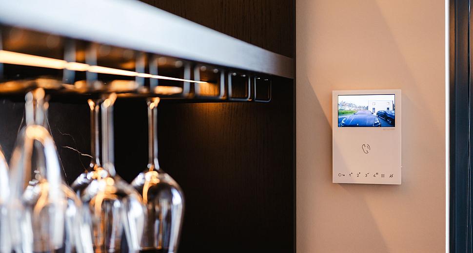 Intercomsysteem met display gemonteerd aan een muur naast een moderne glazenhouder, met zicht op de video-intercominterface die een buitenbeeld toont.
