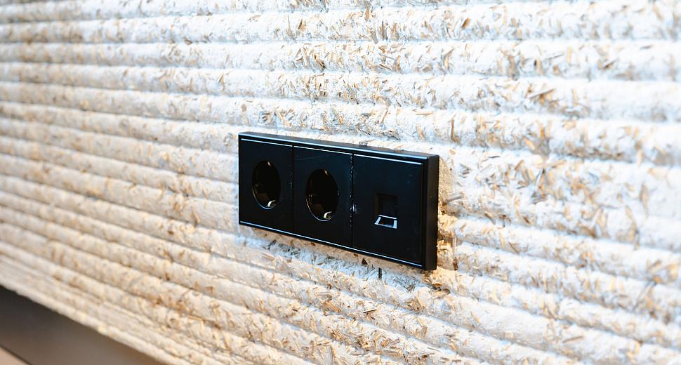 Zwarte wandcontactdoos met twee stopcontacten en een netwerkaansluiting, geplaatst op een decoratieve muur met een gestructureerd beige oppervlak.
