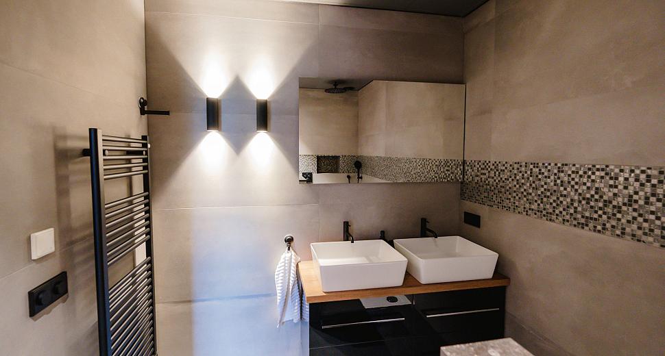 Moderne badkamer met twee witte wastafels op een houten blad, een wandspiegel, een designradiator, grijze tegels en mozaïekdetails op de muur.
