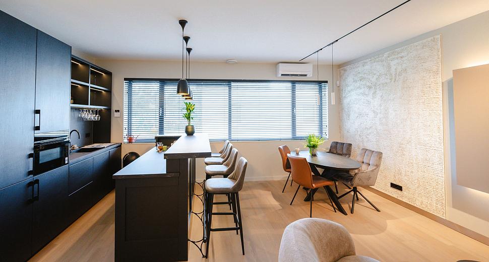Moderne keuken en eetruimte met een zwart kookeiland, barkrukken, eettafel met leren stoelen, grote ramen met jaloezieën en een strakke wandafwerking.