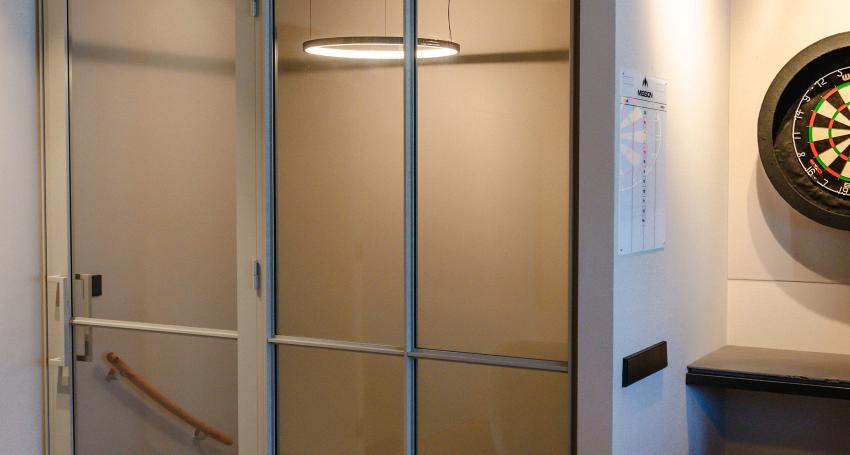 Glazen deur en wand met stalen frame, zicht op een trap met houten leuning en een moderne hanglamp, naast een dartbord aan de muur.