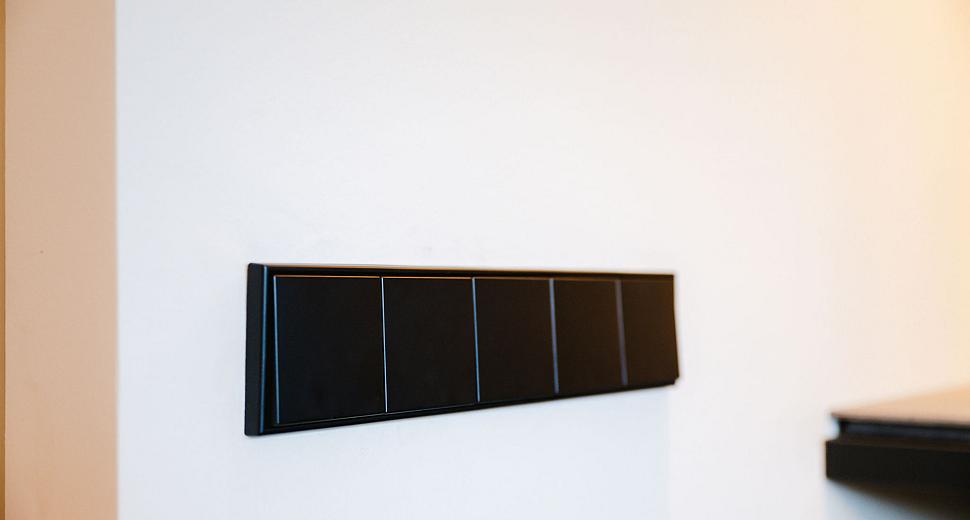 Zwarte moderne lichtschakelaars met strakke lijnen op een witte muur, naast een donkergekleurde plank.