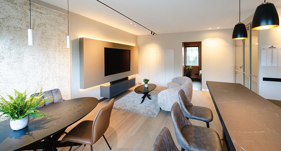 Moderne woonkamer met een strakke inrichting, voorzien van een grote tv aan de muur, een ronde eettafel met stoelen, en stijlvolle hanglampen boven een bar.