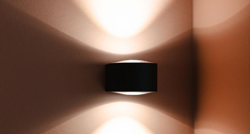 Wandlamp met een zwarte behuizing die zowel omhoog als omlaag licht verspreidt, geplaatst in een hoek van een kamer met zachte, warme verlichtingseffecten.