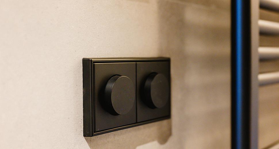 Zwart matte draaiknoppen op een strakke wandplaat, gemonteerd op een beige muur naast een verwarmingsradiator in een moderne badkameromgeving.