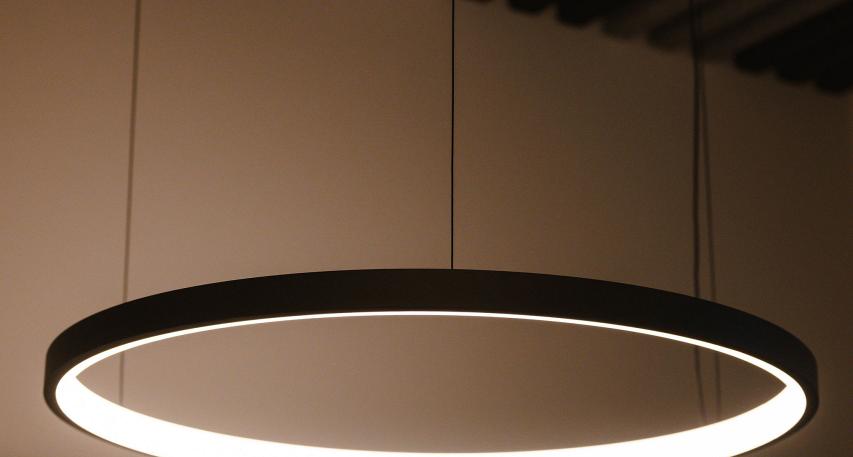  Minimalistische ronde hanglamp met geïntegreerde LED-verlichting, bevestigd aan het plafond met dunne kabels, tegen een achtergrond van houten lamellenplafond.