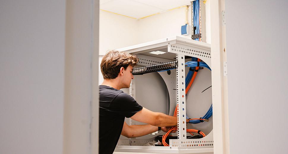 Technicus werkt in een serverkast met oranje en blauwe kabels in een technische ruimte met lichte muren en een systeemplafond.
