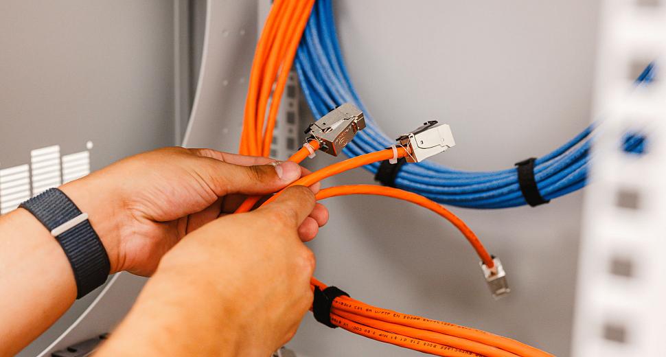 Handen van een technicus die oranje netwerkkabels met metalen connectoren aansluit in een serverkast, met blauwe kabels op de achtergrond.