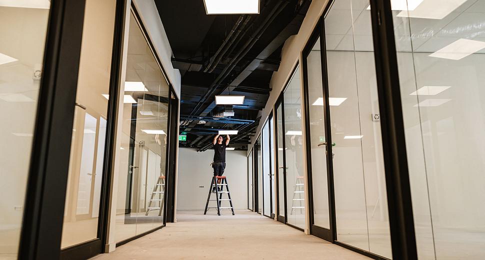 Technicus op een ladder in een moderne gang met glazen wanden, bezig met het installeren van plafondverlichting in een systeemplafond.