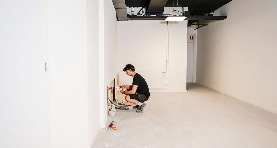Technicus knielt in een lege ruimte en installeert een elektrische aansluiting aan een muur, omringd door kabels en gereedschap, in een onafgewerkte ruimte.