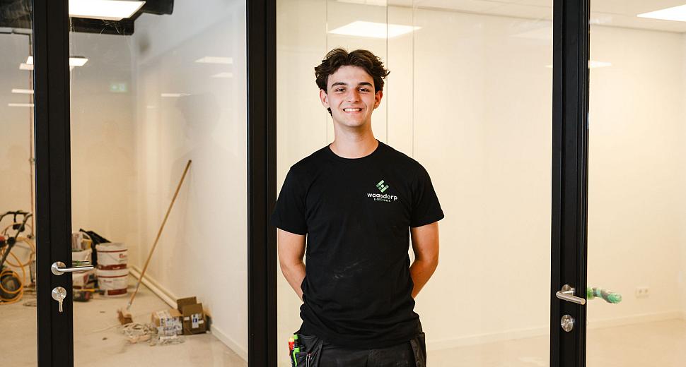 Medewerker in Waasdorp-kleding staat glimlachend in een moderne kantoorruimte met glazen wanden, omringd door bouwmaterialen en gereedschap op de achtergrond.
